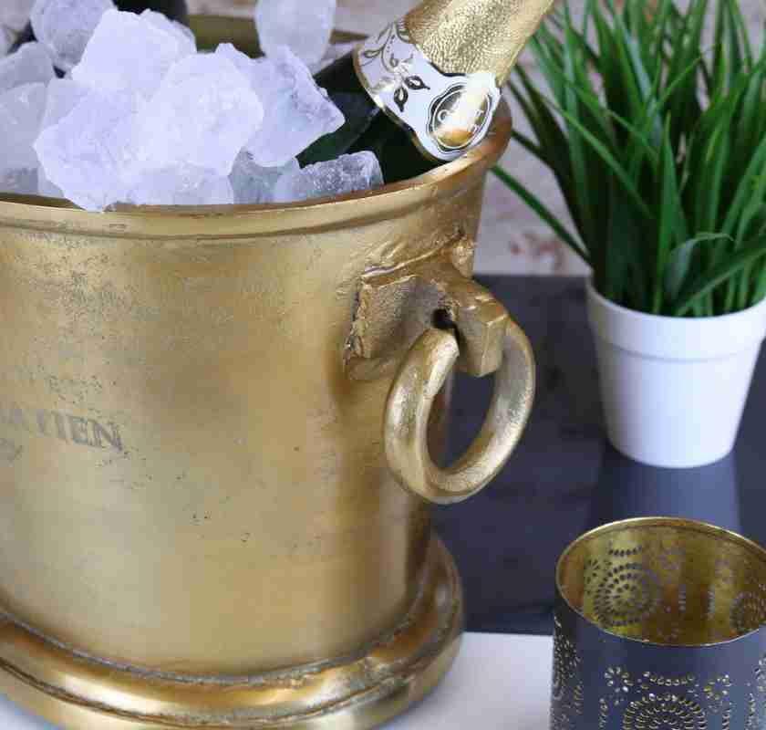 En guldfärgad champagnekylare med is och en flaska champagne. En grön växt och en ljushållare står bredvid.